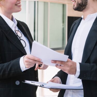 Deux personnes en costume discutent en échangeant des documents.