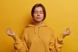 Jeune femme sereine en sweat jaune pratiquant la méditation, yeux fermés dans une pose zen sur fond jaune