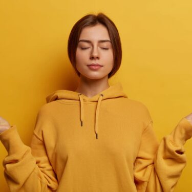 Jeune femme sereine en sweat jaune pratiquant la méditation, yeux fermés dans une pose zen sur fond jaune