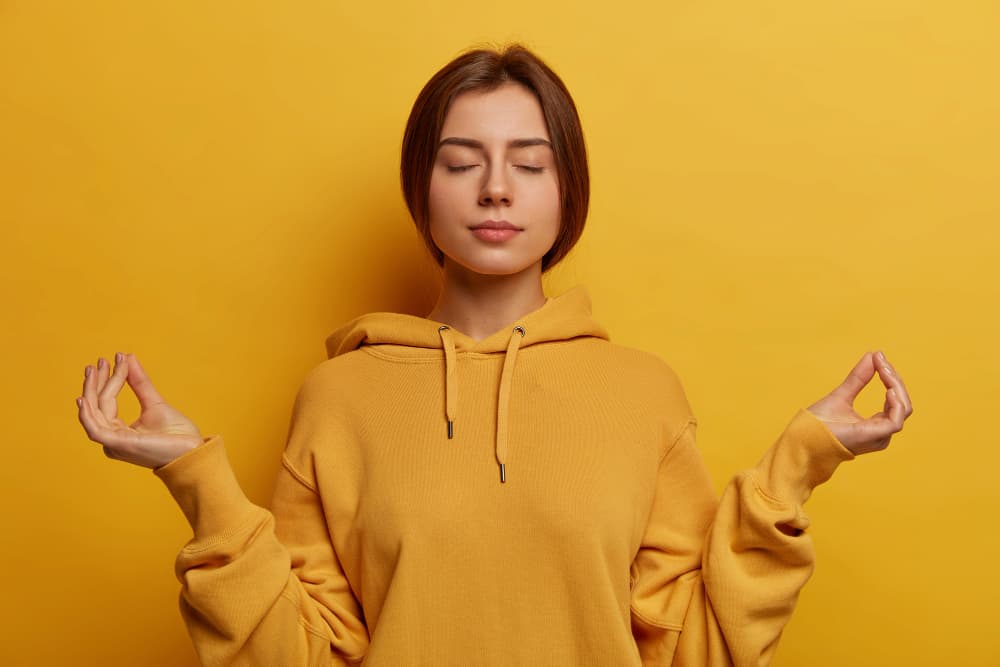 Jeune femme sereine en sweat jaune pratiquant la méditation, yeux fermés dans une pose zen sur fond jaune