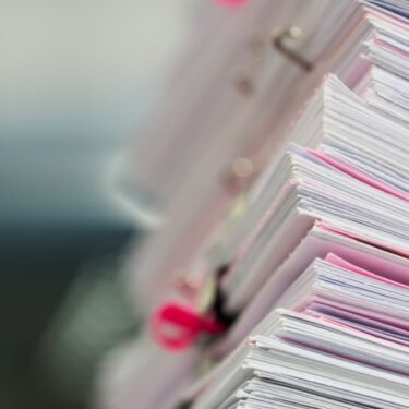 Gros plan sur une pile de dossiers administratifs et réglementaires avec intercalaires roses, illustrant la charge documentaire dans la gestion d'un cabinet