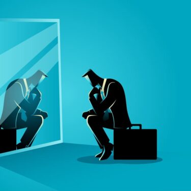 Homme d'affaires assis, pensif, regardant son reflet dans un miroir, sur fond bleu uni.