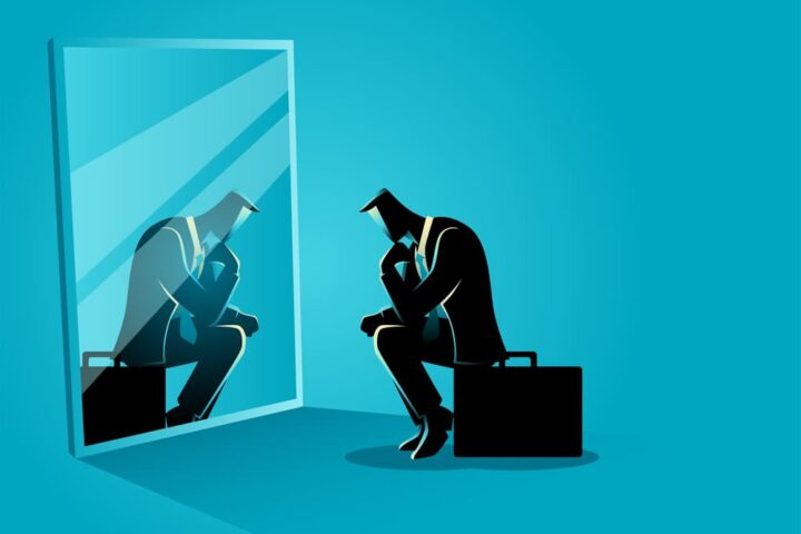 Homme d'affaires assis, pensif, regardant son reflet dans un miroir, sur fond bleu uni.