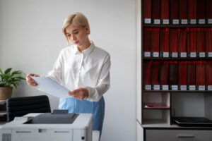 Femme de bureau en chemise blanche examinant des documents près d’une imprimante dans un environnement professionnel.
