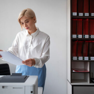 Femme de bureau en chemise blanche examinant des documents près d’une imprimante dans un environnement professionnel.